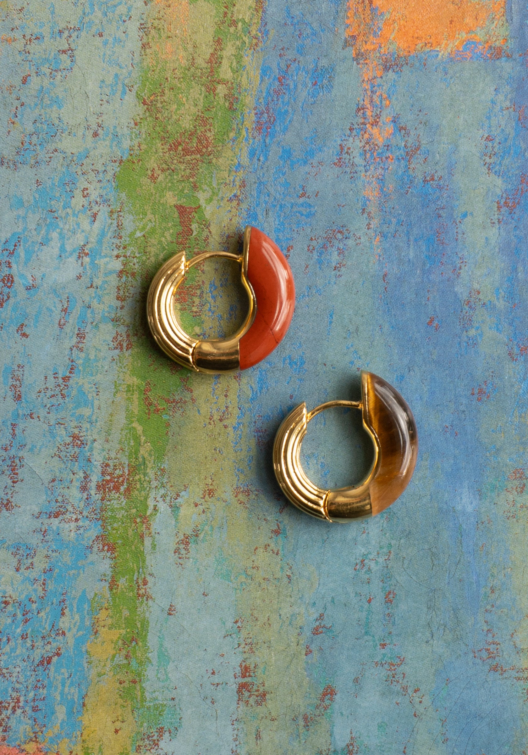 Two gold hoop earrings with red and brown stones on a textured multicolored background